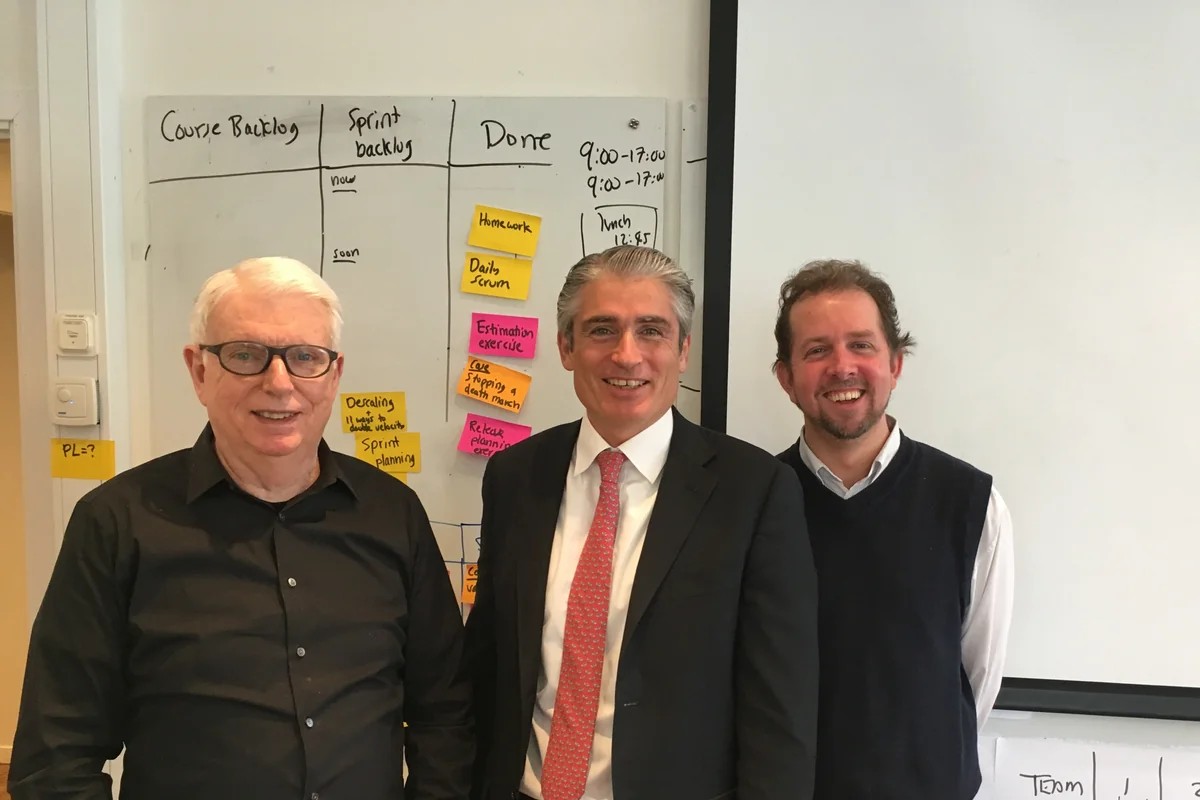 Riccardo Mariti standing between Dr. Jeff Sutherland and Henrik Kniberg in a Scrum training room, a course backlog and sprint board on the whiteboard behind them