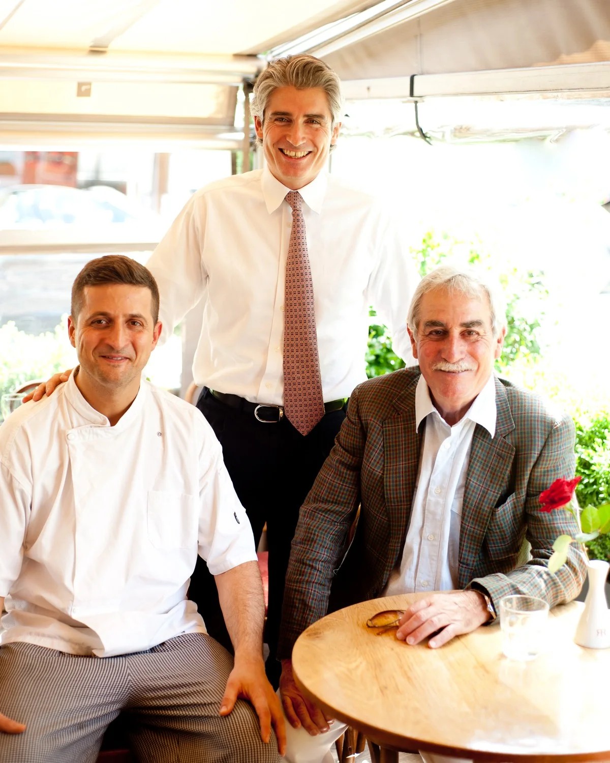 Riccardo Mariti standing behind a seated chef in whites and an older man at a table, on the sunlit terrace at Riccardo's in Chelsea